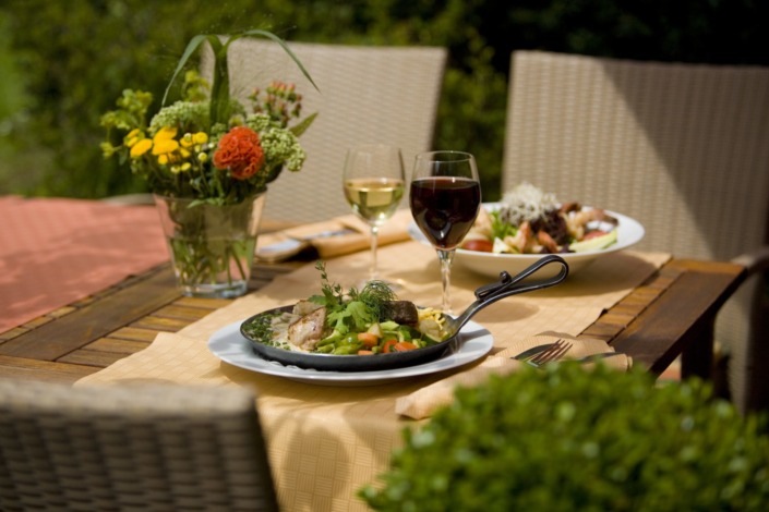 HE_Pfanne+Salat_D9O7078 Zwei Teller mit Essen und zwei Gläser Wein auf einem Esstisch im Freien mit einem Blumenarrangement und Korbstühlen.