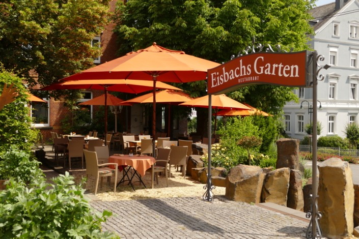 HE_Garten Sitzbereich im Freien eines Restaurants mit orangefarbenen Sonnenschirmen und einem Schild mit der Aufschrift „Eisbachs Garten Restaurant“ an einem sonnigen Tag.