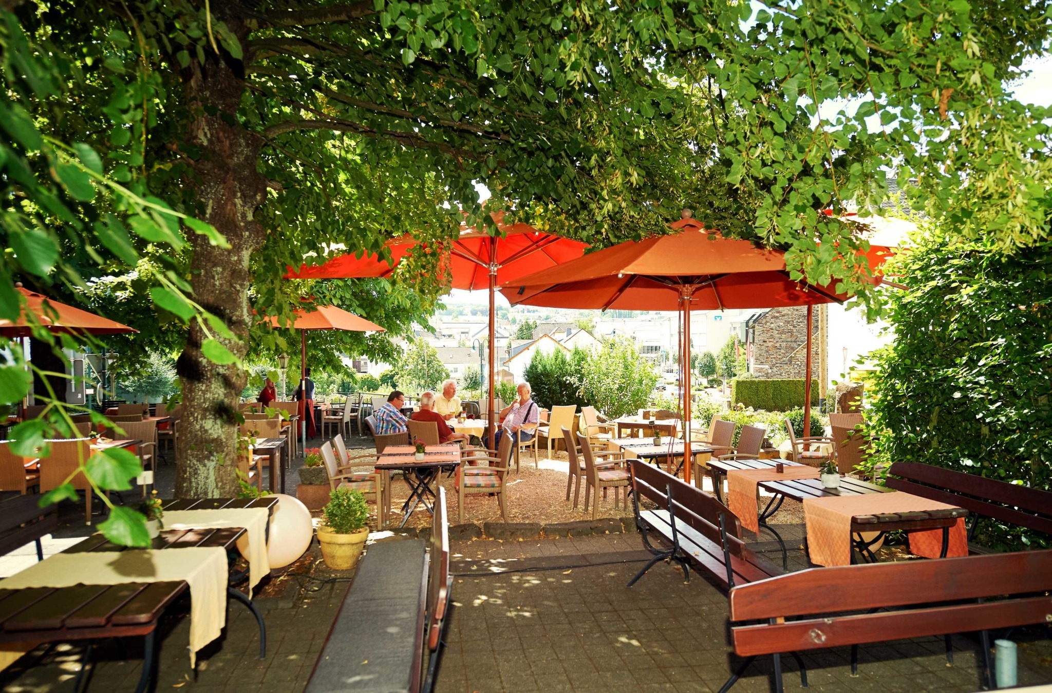 Straßencafé mit Holztischen und Bänken unter großen Bäumen und orangefarbenen Sonnenschirmen, Menschen sitzen und genießen den sonnigen Tag.