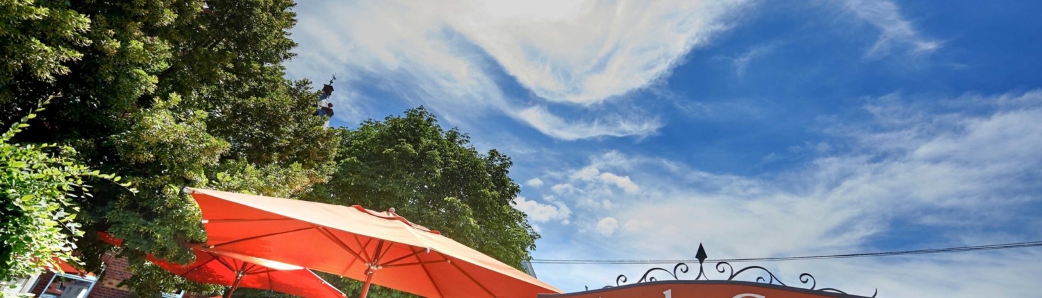 Außenterrasse eines Restaurants mit roten Sonnenschirmen, Tischen und Stühlen unter sonnigem Himmel; Schild „Eisbachs Garten Restaurant“ zu sehen.