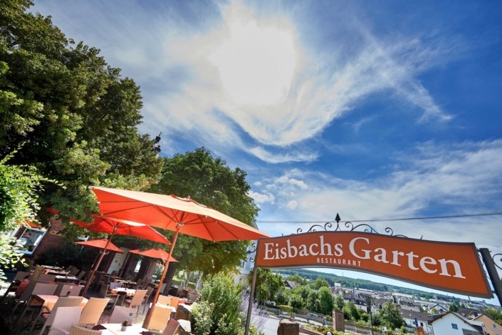 _SG56194 Außenterrasse eines Restaurants mit roten Sonnenschirmen, Tischen und Stühlen unter sonnigem Himmel; Schild „Eisbachs Garten Restaurant“ zu sehen.