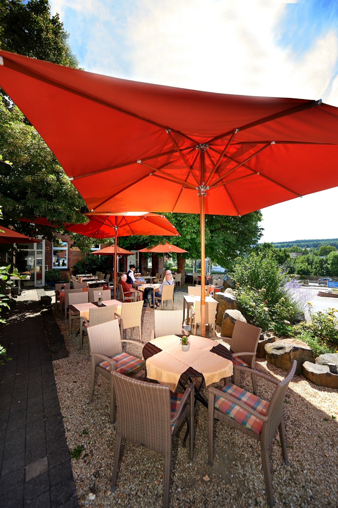 Straßencafé mit Tischen und Korbstühlen unter großen roten Sonnenschirmen, umgeben von Grün und mit Blick auf einen Fluss im Hintergrund.