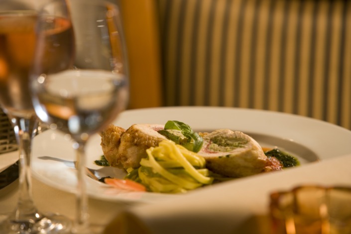 HE_Speisen_HuhnTagliatelle_D9O7085 Ein Gourmet-Menü auf dem Teller mit Hühnchen, Pasta und Beilage, dazu zwei Gläser Wein auf einem Restauranttisch.