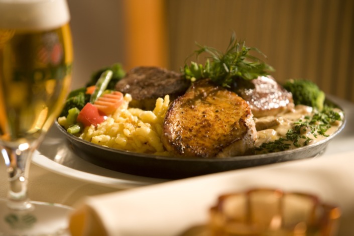 HE_Speisen_Pfännchen_D9O7131 Ein Teller mit gegrilltem Fleisch, Spätzle, Gemüse und Kräutern neben einem Glas Bier auf einem Tisch.
