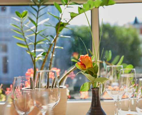 Ein sonnenbeschienener Tisch, gedeckt mit Weingläsern, einer gefalteten Serviette und einer Vase mit einer orangefarbenen Rose, vor einem Fenster mit grünen Pflanzen.