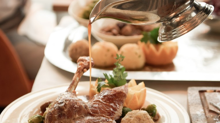 Auf einem weißen Teller an einem Esstisch wird ein gebratenes Fleischgericht mit Knödeln und Gemüse mit Soße übergossen.