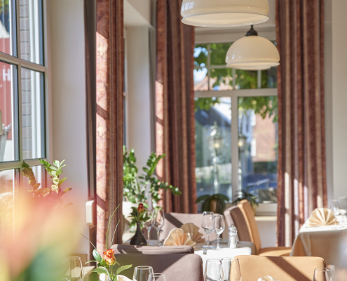 Helles, elegantes Restaurant-Interieur mit gedeckten Tischen, Servietten, Weingläsern und großen Fenstern, die natürliches Sonnenlicht hereinlassen.