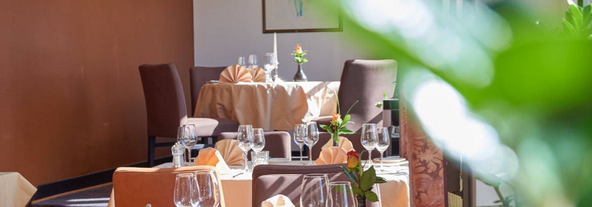 Helles, elegantes Restaurant mit gedeckten Tischen, braunen Stühlen, weißen Tischdecken und Sonnenlicht, das durch große Fenster strömt.