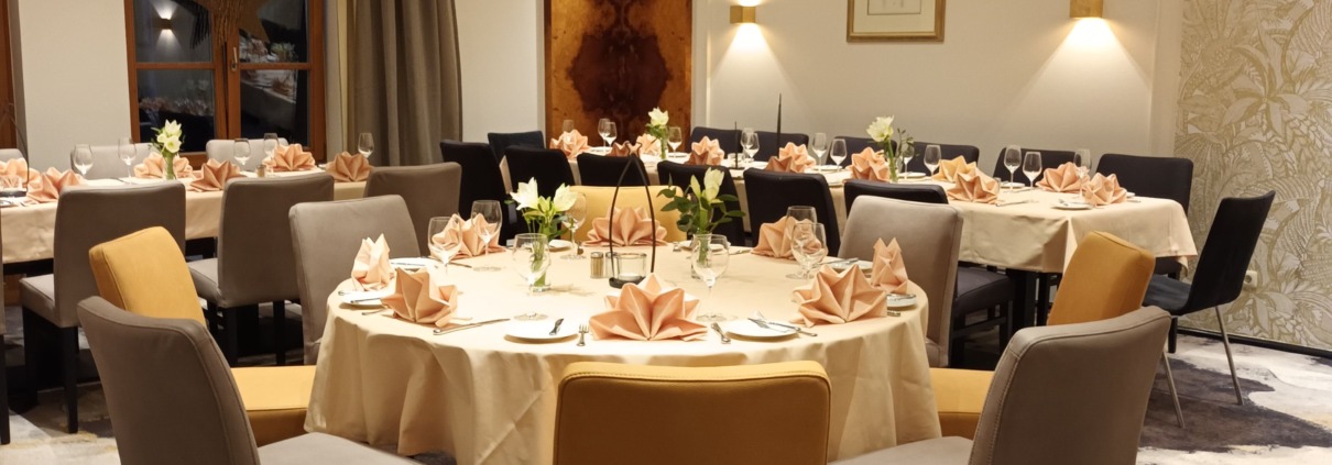 Eleganter Speisesaal eines Restaurants mit runden Tischen, ordentlich gefalteten Servietten und beigen, grauen und goldenen Stühlen, die um jeden Tisch herum angeordnet sind.