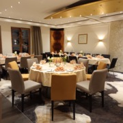 Eleganter Speisesaal eines Restaurants mit runden Tischen, ordentlich gefalteten Servietten und beigen, grauen und goldenen Stühlen, die um jeden Tisch herum angeordnet sind.