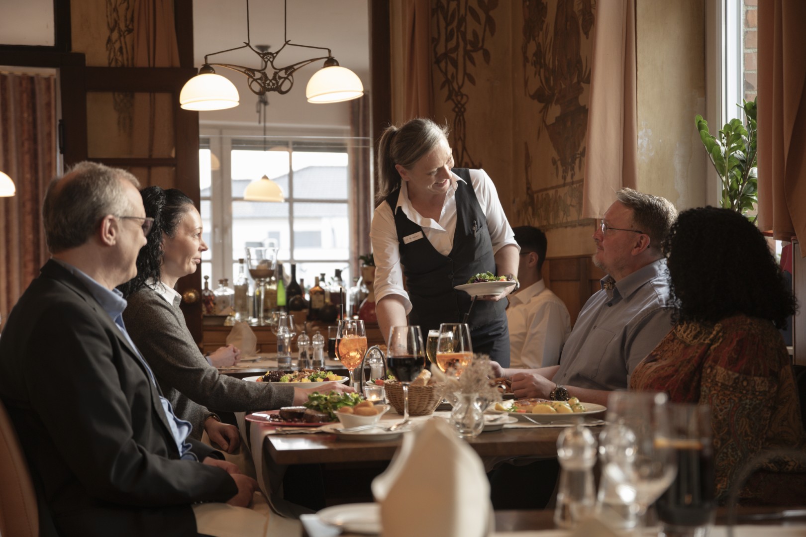 Eine Kellnerin serviert vier Personen, die gemeinsam in einem gemütlichen, warm beleuchteten Restaurant speisen, Essen.
