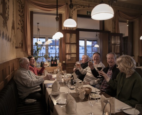 Sechs ältere Erwachsene sitzen an einem Restauranttisch, stoßen an und lächeln, während der Tisch für eine Mahlzeit gedeckt ist.