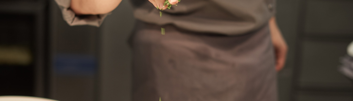 Ein Koch in einer grauen Uniform garniert in einer professionellen Küche ein Tellergericht mit Kräutern.