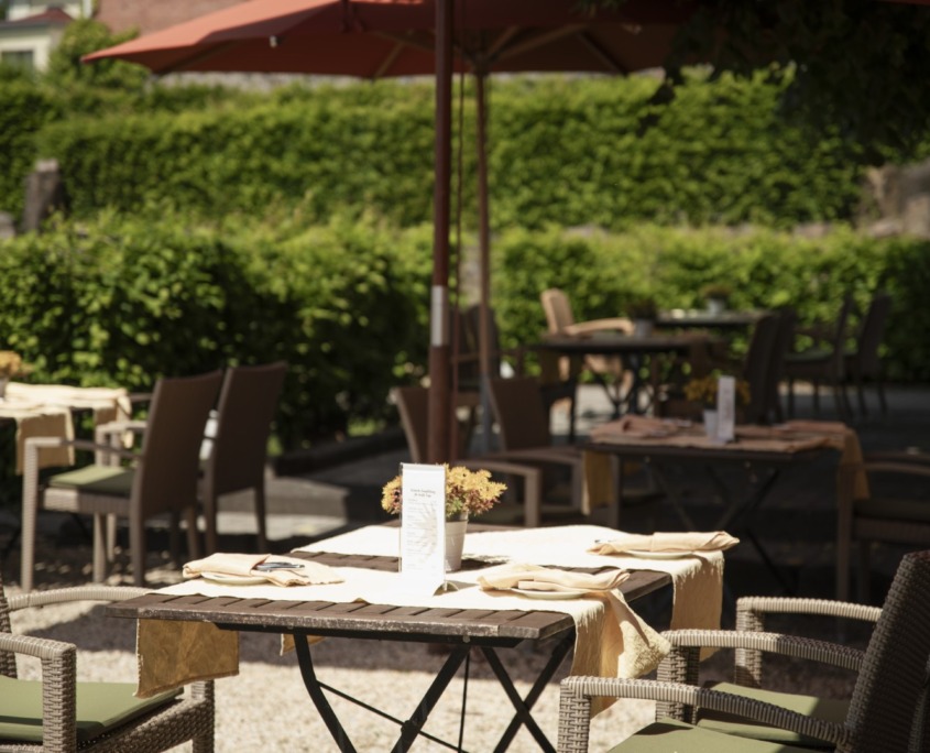_DSC1490 (Groß) Restaurantterrasse mit Korbstühlen, Tischen mit Speisekarten und Blumen sowie großen Sonnenschirmen, die Schatten spenden.