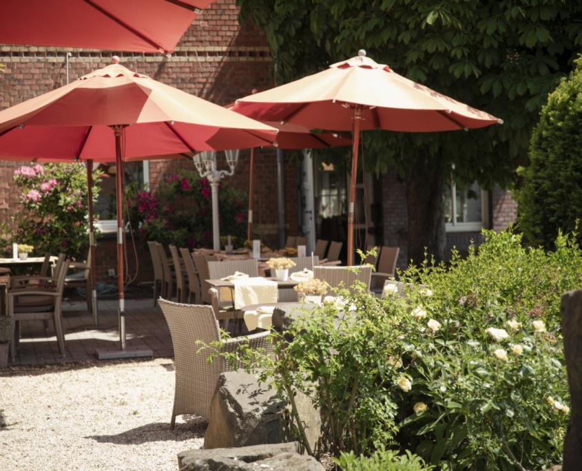 _DSC1495 (Groß) Terrasse im Freien mit Tischen und Stühlen unter großen roten Sonnenschirmen, umgeben von Grün, Blumen und Sonnenlicht.