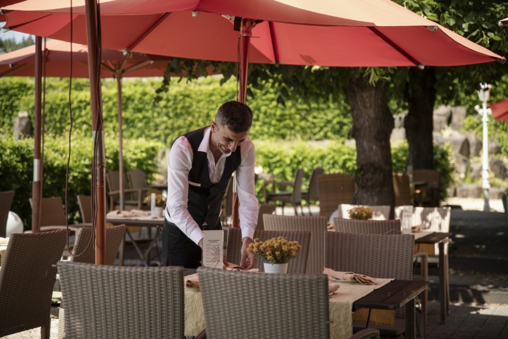 Ein Kellner in weißem Hemd und schwarzer Weste deckt einen Tisch unter roten Sonnenschirmen in einem Außenrestaurant mit leeren Stühlen.