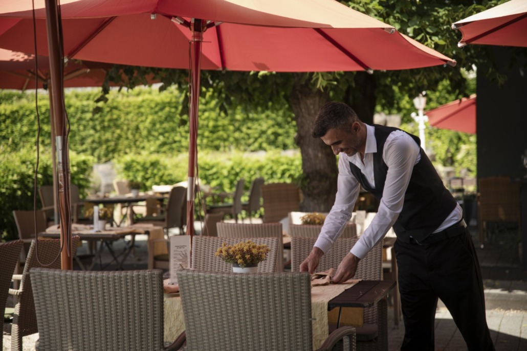 Ein Kellner mit Weste deckt an einem sonnigen Tag einen Tisch in einem Straßencafé unter roten Sonnenschirmen, mit leeren Stühlen und Grünzeug drum herum.