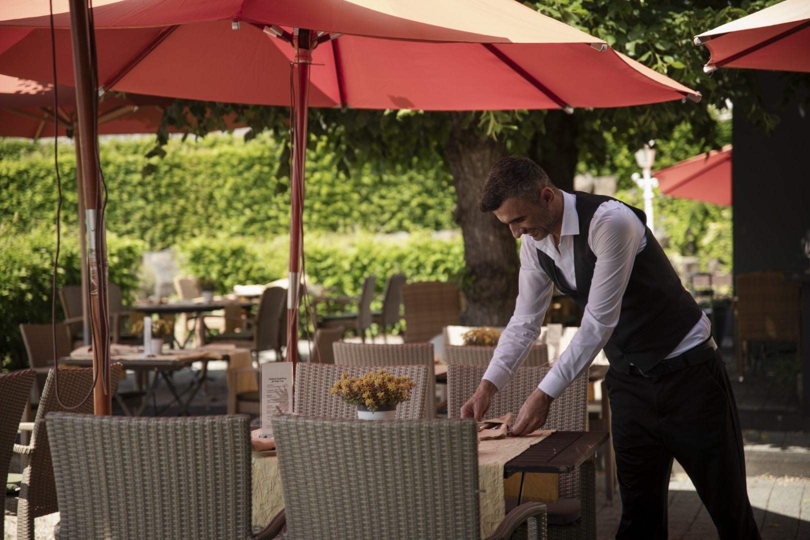Ein Kellner mit Weste deckt an einem sonnigen Tag einen Tisch in einem Straßencafé unter roten Sonnenschirmen, mit leeren Stühlen und Grünzeug drum herum.