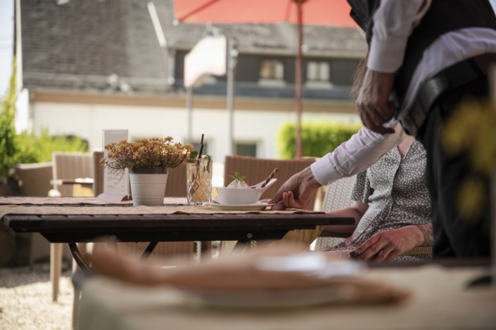 _DSC1660 (Groß) Ein Kellner stellt eine Schale für einen sitzenden Gast auf den Tisch eines Straßencafés; ein Getränk und eine Topfpflanze stehen ebenfalls auf dem Tisch.
