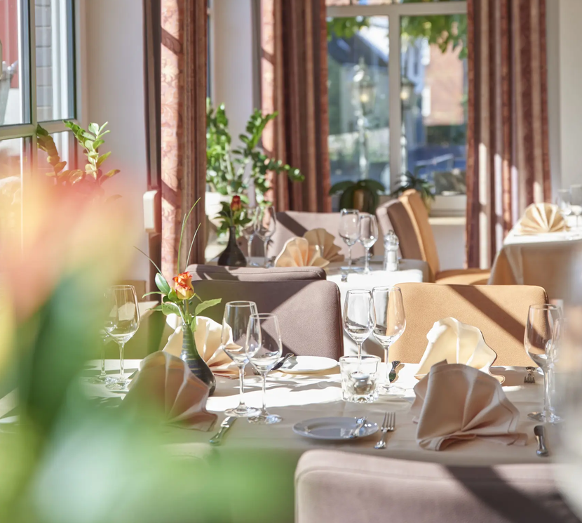 Restauranttische, gedeckt mit weißen Tischdecken, gefalteten Servietten und Gläsern, in einem sonnendurchfluteten Raum.